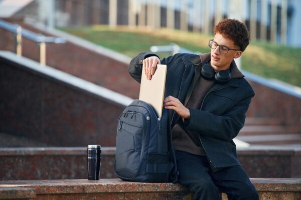 Taking out laptop from backpack. Young student is outside the university, in black clothes